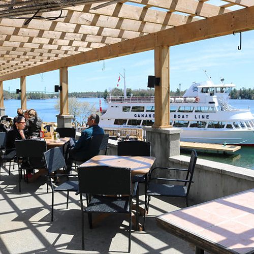 A group of people sit beneath a pergola as a tour boat sails in the background.