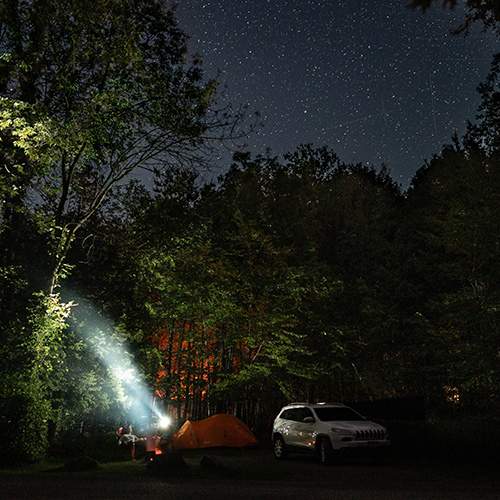 A white SUV sits beside an orange tent underneath a canopy of trees at night, with a starry sky.